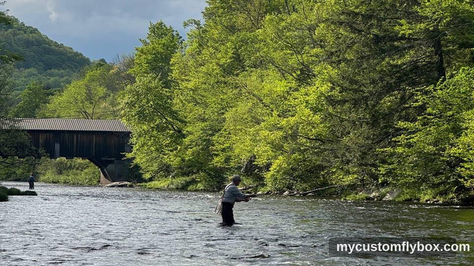 Angler fly fishing a Catskills river near a covered bridge under a cloudy spring sky.